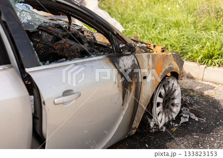 A car stands at the roadside, severely damaged by fire. Its body features burnt areas and a destroyed interior, with debris and ash around, reflecting a recent incident of a vehicle fire 133823153