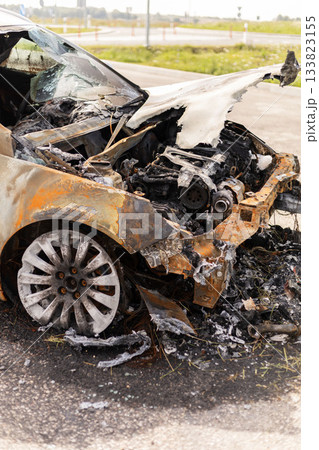 A burned car sits on the roadside, completely charred and damaged. The vehicle shows signs of extensive fire damage, with melted parts and a blackened body, indicating a serious fire incident 133823155