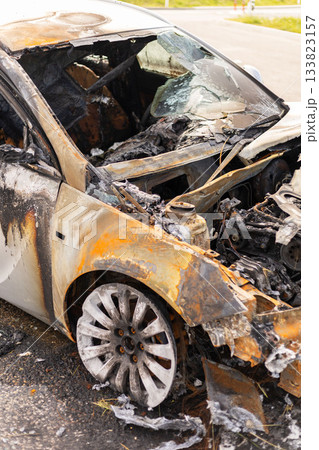 Burned car wreckage displays extensive damage with charred metal and ash. The vehicle rests by the road in an industrial area, showing signs of a recent fire Burned car wreckage displays extensive damage with charred metal and ash. The vehicle rests by the road in an industrial area, showing signs of a recent fire 133823157