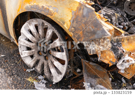 A burned-out vehicle sits in an empty lot, with a damaged wheel and charred exterior visible. Scattered debris and remnants of the fire surround the wreck, revealing the extent of the damage 133823159