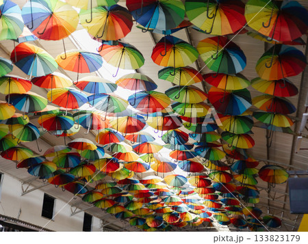 A multitude of colorful umbrellas is creatively arranged overhead in a bustling market. The bright hues add a cheerful atmosphere, drawing visitors' attention A multitude of colorful umbrellas is creatively arranged overhead in a bustling market. The bright hues add a cheerful atmosphere, drawing visitors' attention 133823179