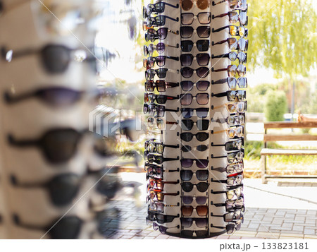 A vibrant outdoor market scene features a tall display of sunglasses. Different styles and colors are arranged neatly, inviting shoppers to browse. The sunlight adds a warm touch to the setting 133823181