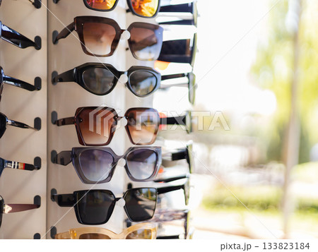 A selection of stylish sunglasses hangs on a display rack. The sun shines brightly, highlighting the different shapes and colors. The setting suggests a lively shopping atmosphere in summer 133823184