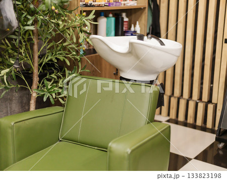 A vibrant green salon chair is positioned beside a sleek hair washing station. The setting is stylish, with wooden shelves displaying hair products, creating a modern and inviting atmosphere 133823198