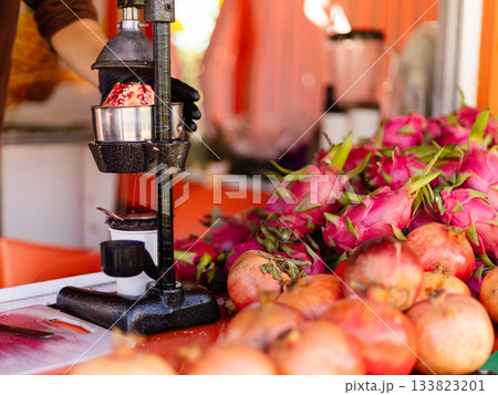 A fruit vendor uses a juicer to extract juice from bright pomegranates while surrounded by colorful dragon fruit. The cheerful setting enhances the freshness of the offerings 133823201
