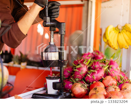 A vendor presses ripe pomegranates to make fresh juice at a vibrant market stand. Colorful fruits, including dragon fruit and bananas, are displayed nearby 133823204