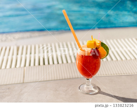 A colorful tropical cocktail is placed on a stone surface next to a pool. The drink features vibrant garnishes and a straw, perfect for enjoying under the sun. Copy space. 133823216