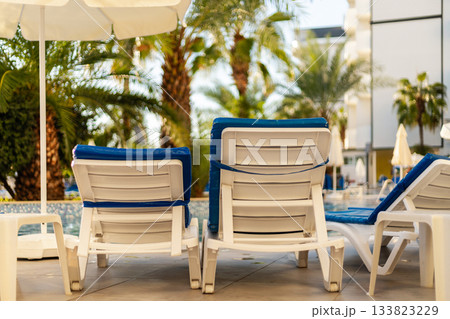 Comfortable white lounge chairs with blue cushions sit near a sparkling pool, surrounded by lush palm trees under a bright sun. Guests can enjoy a relaxing day outdoors. All inclusive. 133823229