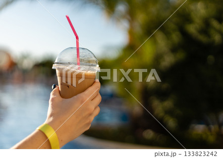 A hand holds a clear cup of iced coffee with a pink straw, set against a sunny backdrop of palm trees and a sparkling pool. Perfect for a warm day 133823242