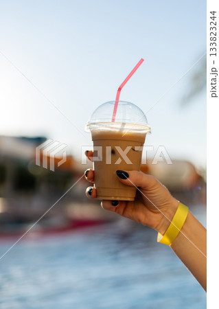 A hand holds a clear cup of iced coffee with a pink straw, set against the backdrop of a bright pool area. The scene captures a relaxing summer moment. All inclusive. 133823244