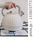 Vertical view a person is holding a white tea kettle over a gas stove 133823773