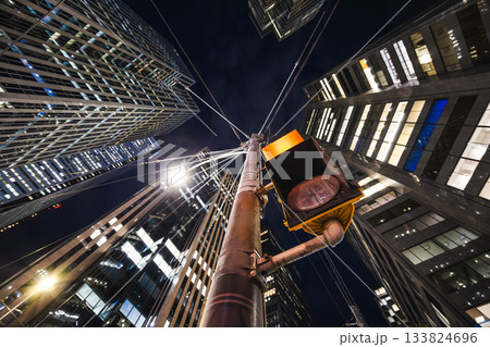 Nighttime upward view of towering glass buildings converging above a glowing traffic light, creating a dynamic sense of motion Nighttime upward view of towering glass buildings converging above a glowing traffic light, creating a dynamic sense of motion 133824696