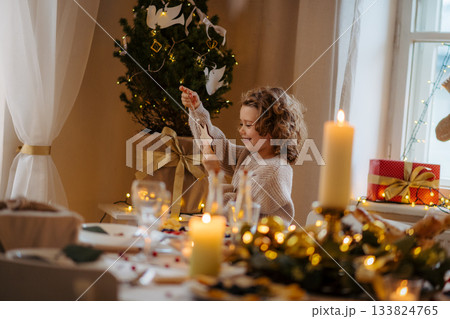 Cheerful small girl indoors at home at Christmas, looking at christmas ornament. Cheerful small girl indoors at home at Christmas, looking at christmas ornament. 133824765