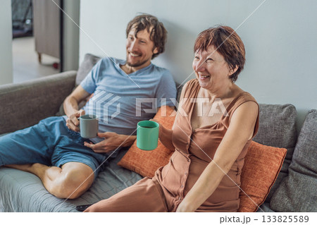 Older woman and her adult son relaxing together on the sofa while watching a comedy on television, enjoying a warm family moment at home. Entertainment, bonding and cozy lifestyle concept 133825589