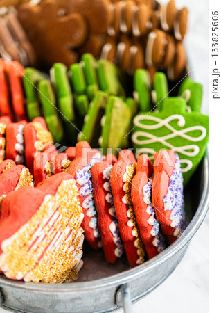 Captured from a close-up perspective, a tray of vibrant holiday cookies is neatly arranged in rows, featuring red and green iced cookies with intricate piping and sandwich-style gingerbread cookies 133825606