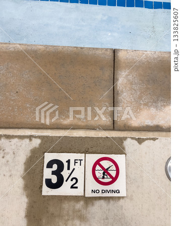A close-up shot of a pool's concrete edge showing the depth marking and safety icon. The image captures the transitional space from deck to water. A close-up shot of a pool's concrete edge showing the depth marking and safety icon. The image captures the transitional space from deck to water. 133825607