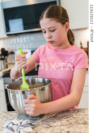 A girl in a pink t-shirt stirs a bowl of cookie dough using a green spatula. She is standing at a kitchen counter with granite surfaces and white cabinetry. 133825633
