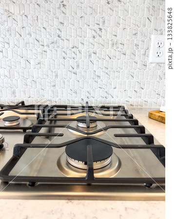 A gas cooktop is positioned in front of a white stone tile backsplash with a power outlet nearby. The image captures a clean and modern culinary space. 133825648