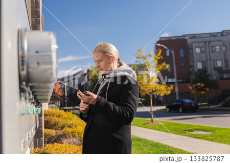 Middle-aged woman checking electricity meters outside a residential building and entering usage data on her smartphone during a sunny autumn day 133825787