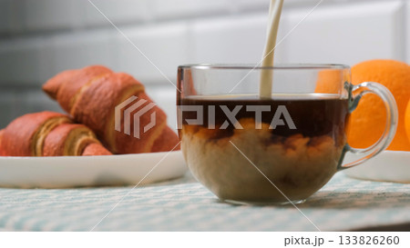 Close-up of milk pouring into a cup with hot coffee, croissants and oranges nearby. Cozy morning comfort concept  133826260