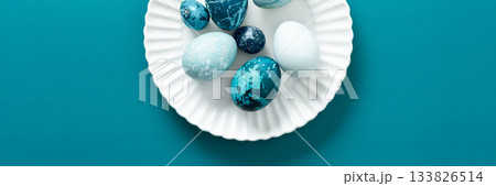 Colorful blue Easter eggs are artistically arranged on a white plate resting on a vibrant teal surface, capturing the essence of spring festivities and holiday traditions, banner 133826514