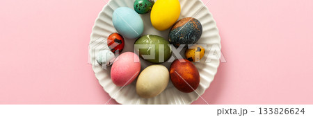 A collection of vibrant Easter eggs in various colors is artfully arranged on a white plate, set against a soft pink background, capturing the essence of the holiday's decorations, banner 133826624