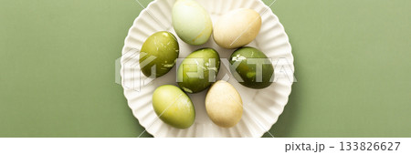 Beautifully decorated eggs in shades of green and cream are artistically arranged on a white plate, perfect for celebrating Easter. The soft green background enhances the festive mood, banner Beautifully decorated eggs in shades of green and cream are artistically arranged on a white plate, perfect for celebrating Easter. The soft green background enhances the festive mood, banner 133826627