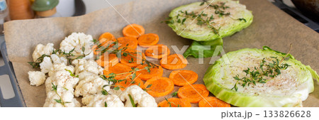 Cauliflower, carrots, and cabbage are sliced and arranged on a tray lined with parchment paper, ready for roasting with herbs in a warm kitchen setting, banner 133826628