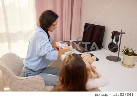 Mother working on a laptop while daughter plays with a teddy bear at home 133827841