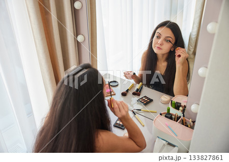 Woman applying mascara to her eyelashes in front of a makeup mirror Woman applying mascara to her eyelashes in front of a makeup mirror 133827861