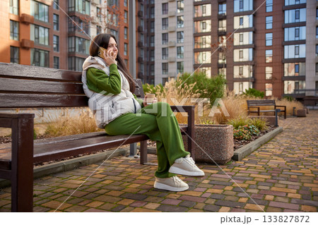 Woman having an outdoor phone conversation while sitting on a bench with coffee Woman having an outdoor phone conversation while sitting on a bench with coffee 133827872
