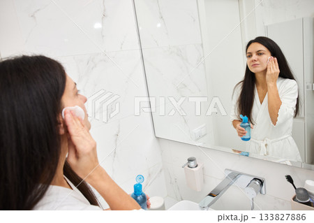 Woman cleansing her face with toner and cotton pad looking in bathroom mirror Woman cleansing her face with toner and cotton pad looking in bathroom mirror 133827886