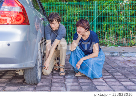 Young man helping an older woman by changing the car wheel while she sits nearby, showing support, kindness and real-life roadside assistance. Family care, safety, teamwork and everyday problem Young man helping an older woman by changing the car wheel while she sits nearby, showing support, kindness and real-life roadside assistance. Family care, safety, teamwork and everyday problem 133828016