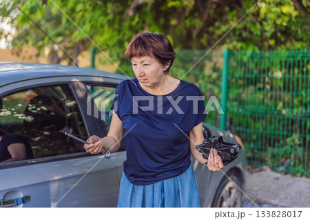 Older woman standing outdoors holding a car jack and looking confused, unsure how to use it. Real-life moment, problem solving and everyday lifestyle concept, showing vulnerability, learning and Older woman standing outdoors holding a car jack and looking confused, unsure how to use it. Real-life moment, problem solving and everyday lifestyle concept, showing vulnerability, learning and 133828017