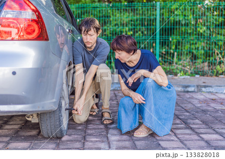 Young man helping an older woman by changing the car wheel while she sits nearby, showing support, kindness and real-life roadside assistance. Family care, safety, teamwork and everyday problem 133828018