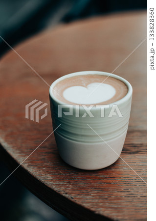 A beautiful cappuccino cup with latte art in a heart shape sits on a rustic wooden table. The warm tones create a cozy feel. A heart-shaped milky foam 133828360