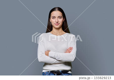 Beautiful young woman with crossed arms looking at camera on grey background Beautiful young woman with crossed arms looking at camera on grey background 133829213