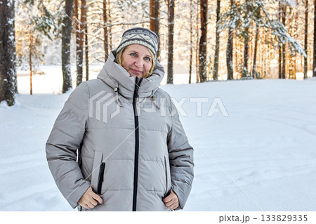 Warmly dressed mature woman stands in snowy forest while her companion takes photo. Warmly dressed mature woman stands in snowy forest while her companion takes photo. 133829335