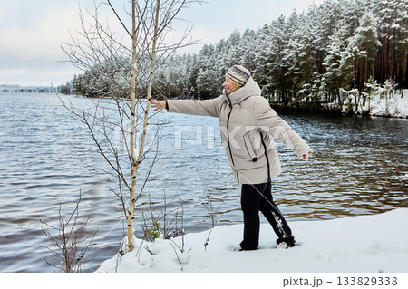 Smiling woman approaches lake edge, reaching for birch branch for support on slippery ground while wearing winter clothes, snowy forest behind. Smiling woman approaches lake edge, reaching for birch branch for support on slippery ground while wearing winter clothes, snowy forest behind. 133829338