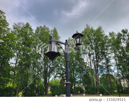 A broken lantern stands against a gloomy sky and a green park. 133829521