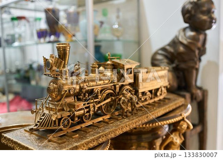 Old brass locomotive on showcase, Historical brass train among shelves and decorative figures 133830007