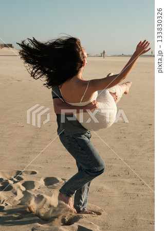 Man carrying woman in his arms on sandy beach, her hair flowing in the wind during a romantic and playful moment. Man carrying woman in his arms on sandy beach, her hair flowing in the wind during a romantic and playful moment. 133830326
