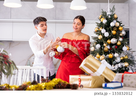 Excited young couple enjoying decorating Christmas tree together while standing in front of wrapped presents in living room 133832012