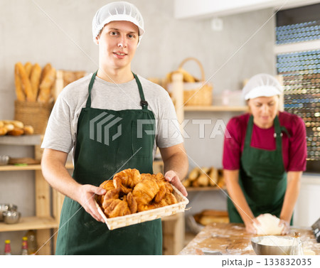 Young baker in uniform stands in small private bakery offering hot croissants 133832035