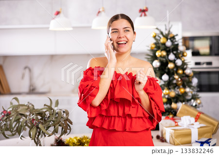 Emotional young woman in red dress inviting guests by calling on phone next to wrapped gifts and Christmas tree in living room at home Emotional young woman in red dress inviting guests by calling on phone next to wrapped gifts and Christmas tree in living room at home 133832246