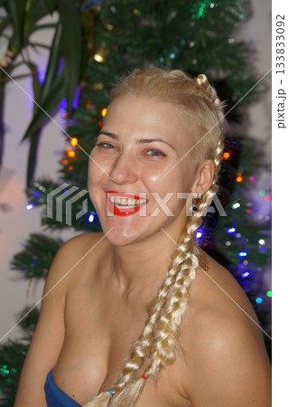 A young blonde looks at the camera, smiles, laughs, and rejoices. In the background is a New Year's tree and garland. Portrait photography. 133833092