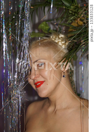 A young blonde model looks at the camera, smiles, laughs, and rejoices. In the background is a New Year's tree and garland. Portrait photography. 133833128