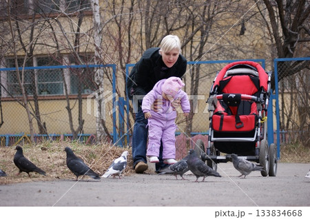 A young woman, a mother, is walking with her child on the street, feeding pigeons. A red stroller is standing nearby. 133834668