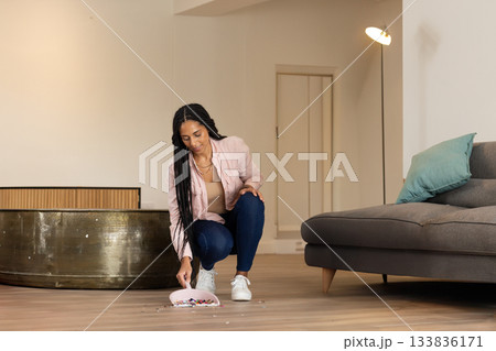 African American woman crouching in living room gathering beads with pink dustpan and brush 133836171
