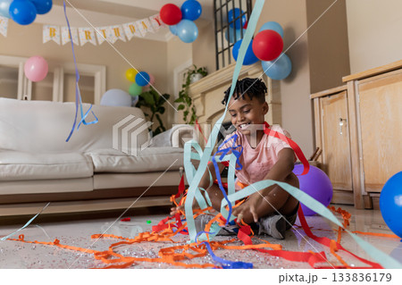 African American boy sitting on floor tossing streamers and balloons in living room, copy space 133836179
